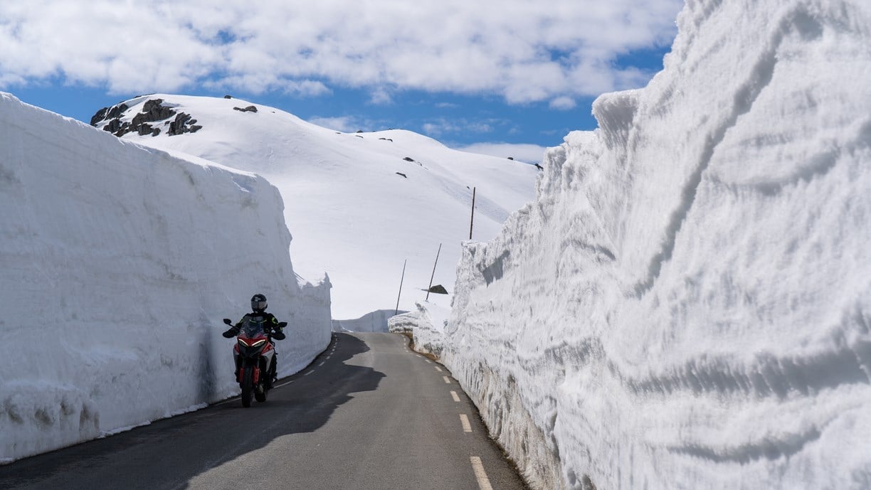 Mye snø over fjellet. Motorsyklist kjører på bar veg med to til tre meter høye brøytekanter.