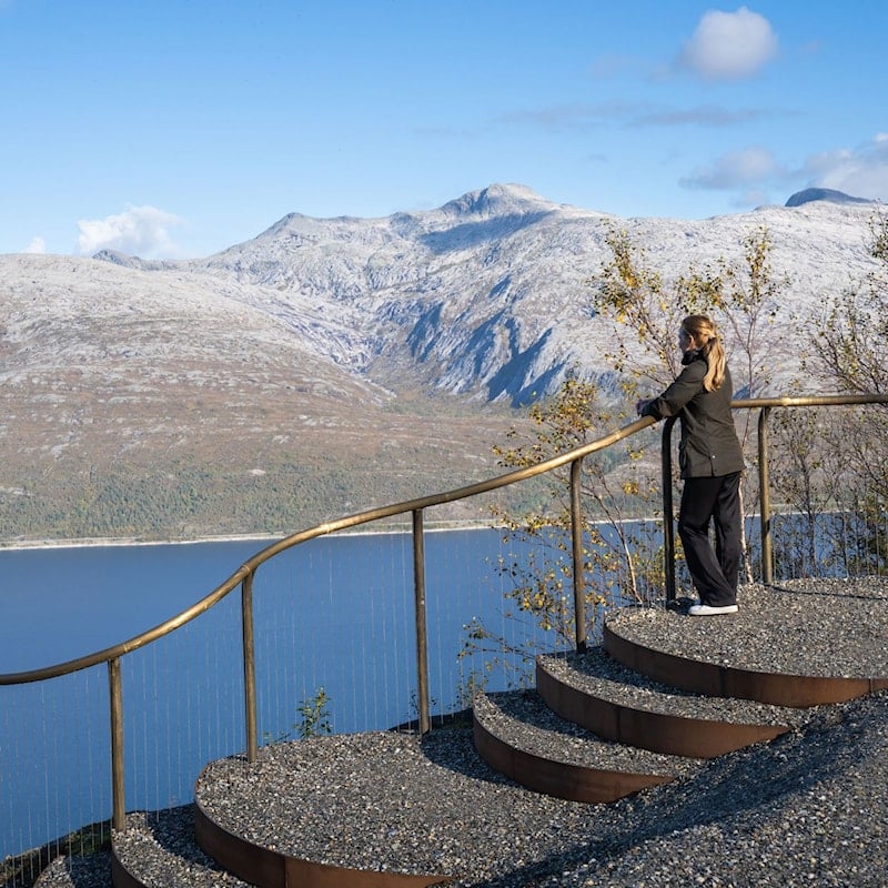 Person på utsiktspunkt med buede terrasser og utsyn over fjord og fjell.