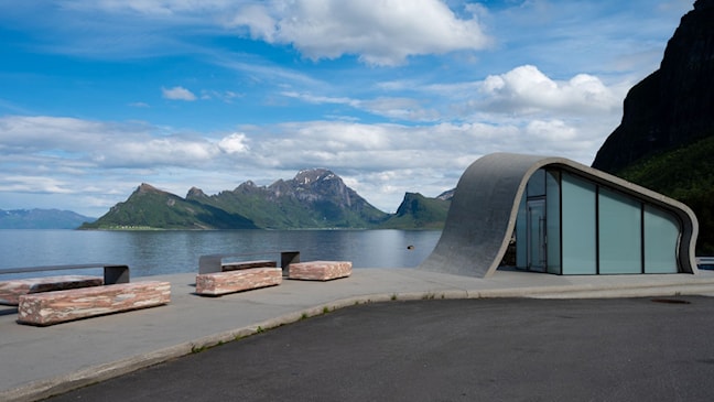 Rasteplass med et buet toalettbygg og rastebenker formet i stein. Utsikt til havet. 