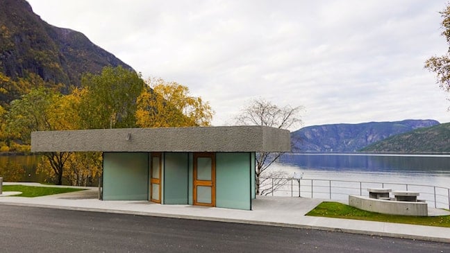 Toalettbygg i betong og glas på Røyrnes ved Lærdalsfjorden. Framfor bygget ligg asfalt og gangareal, medan fjorden, fjell og haustfarga tre dannar bakgrunnen.