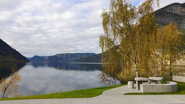 Betongbenkar og bord ved Lærdalsfjorden på Røyrnes. I bakgrunnen ligg fjorden omgjeven av fjell og haustfarga bjørketre.