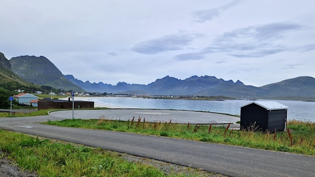 Oversiktsbilde av en rund asfaltert parkeringsplass. En liten toalettbygning står ved siden av. Hav og fjell i horisonten bak. 