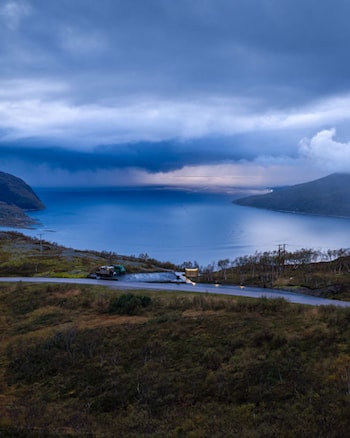 En person går langs en gangvei ved en parkeringsplass med utsikt over fjord og fjell. I forgrunnen står en rund bygning med vertikale trespiler, mens hav og kystlandskap strekker seg ut i bakgrunnen.