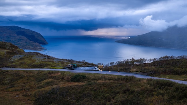 En person går langs en gangvei ved en parkeringsplass med utsikt over fjord og fjell. I forgrunnen står en rund bygning med vertikale trespiler, mens hav og kystlandskap strekker seg ut i bakgrunnen.