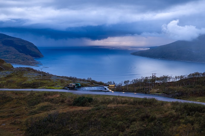 En person går langs en gangvei ved en parkeringsplass med utsikt over fjord og fjell. I forgrunnen står en rund bygning med vertikale trespiler, mens hav og kystlandskap strekker seg ut i bakgrunnen.