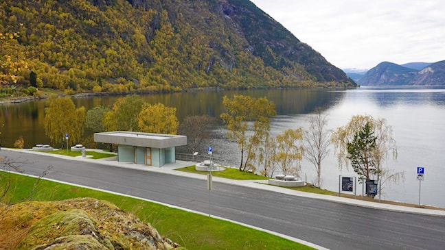 Moderne toalettbygg ved fjorden. Foran bygget er det gangareal og sitjeplassar, med bratte fjellsider og Lærdalsfjorden i bakgrunnen.