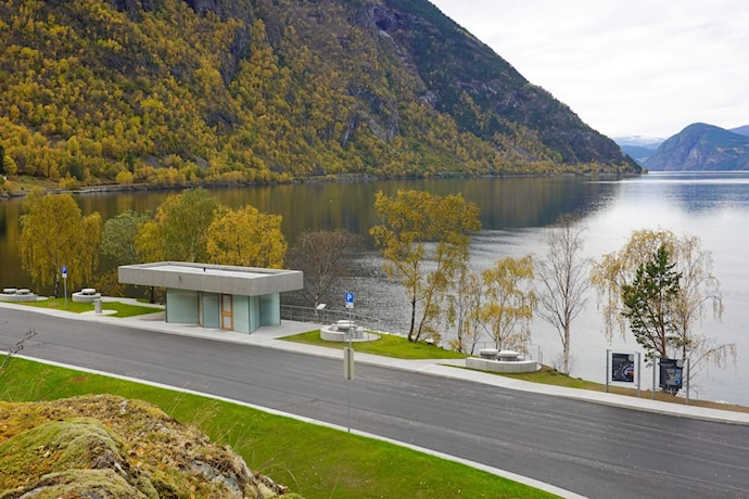 Moderne toalettbygg ved fjorden. Foran bygget er det gangareal og sitjeplassar, med bratte fjellsider og Lærdalsfjorden i bakgrunnen.
