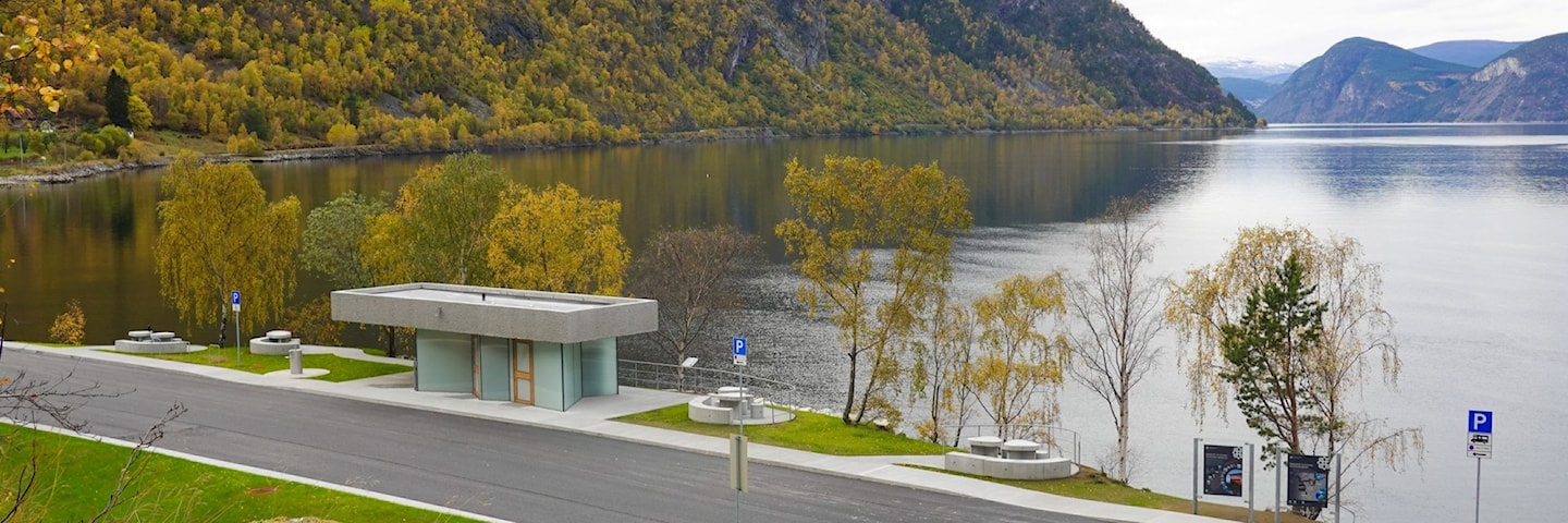 Moderne toalettbygg ved fjorden. Foran bygget er det gangareal og sitjeplassar, med bratte fjellsider og Lærdalsfjorden i bakgrunnen.