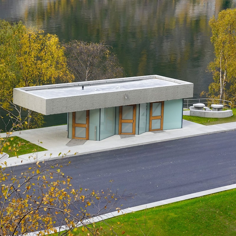 Toalettbygg og sitjeområde ved Røyrnes rasteplass, omgitt av tre, grøntareal og fjell.