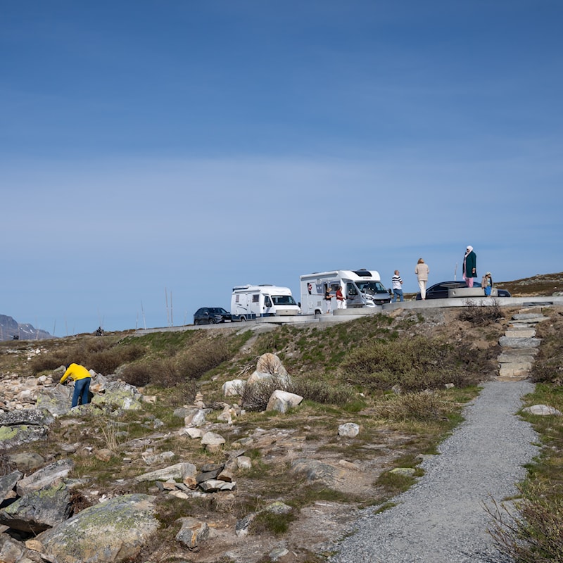 Bilde av en gruset sti. I bakgrunnen parkerte biler og personer som tar en pause.